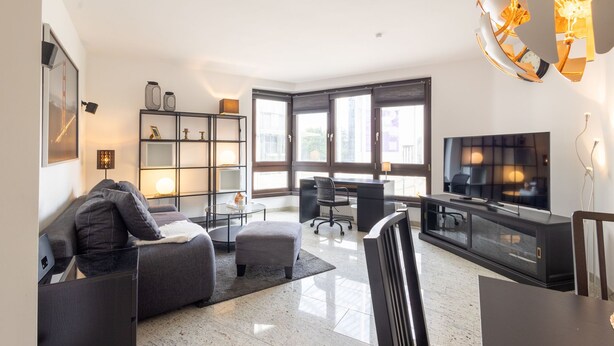 A modern living room with a sofa, coffee table, and TV stand. There is a bookshelf and a desk with a chair. Large windows provide natural light.
