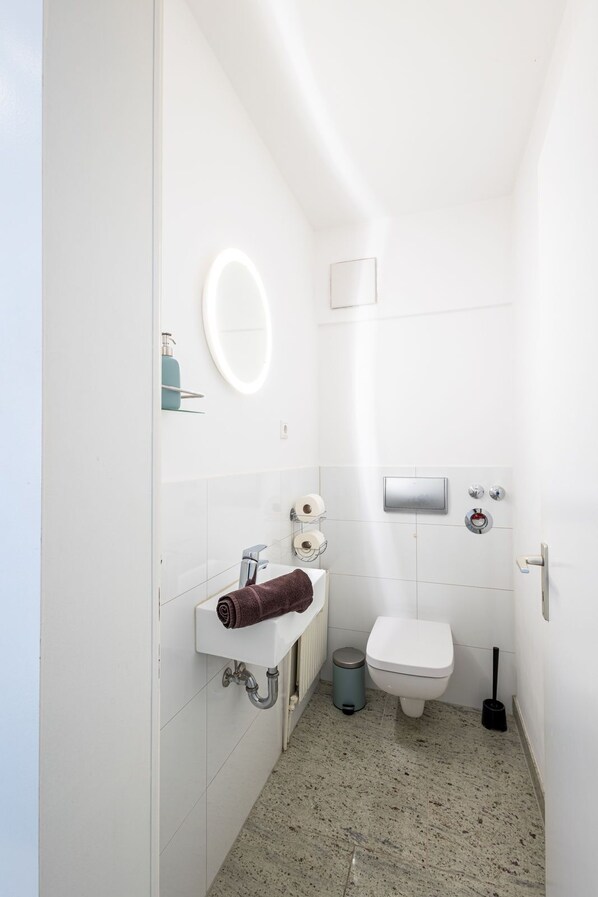 A small, white bathroom with a sink, mirror, toilet, and a trash bin.