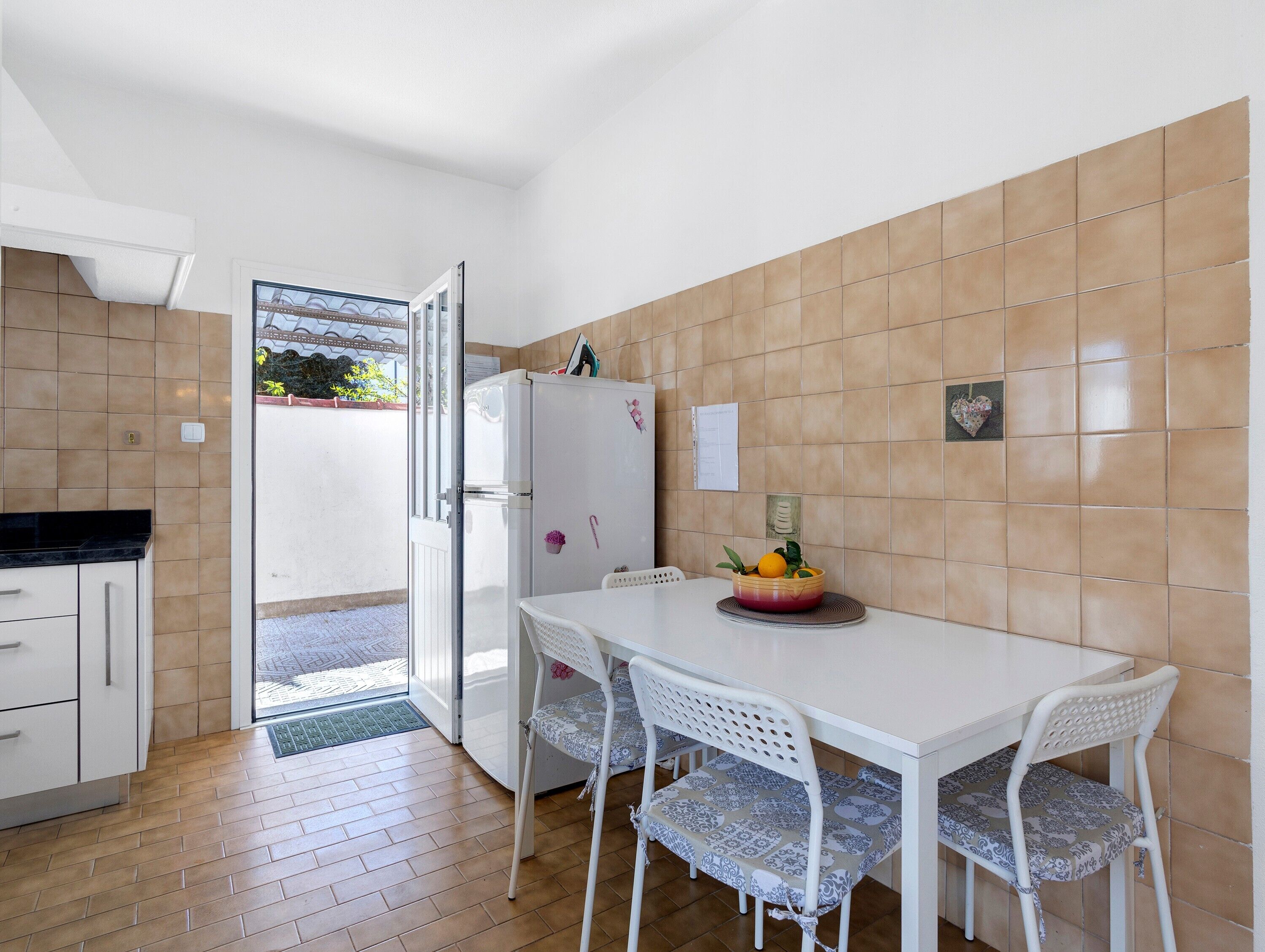 Une cuisine compacte avec un réfrigérateur blanc, une table à manger avec deux chaises, et une vue sur un espace extérieur accessible par une porte ouverte.