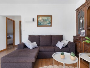 Un salon comprenant un canapé d’angle, une petite table ronde agrémentée de deux plantes en pot, un meuble en bois et un tableau accroché au mur.