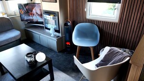 Ein kompakter Wohnraum mit Fernseher, Heizung, blauem Sessel, weißem Stuhl und einem kleinen Tisch mit Tasse.