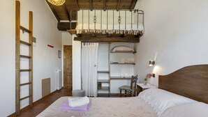 Un dormitorio con cama alta, una escalera, una silla, una mesita y una repisa.