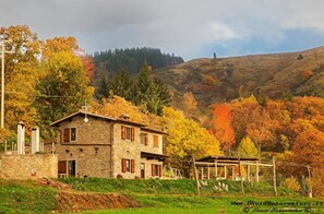 Una casa de piedra con balcón, rodeada de follaje otoñal y colinas.