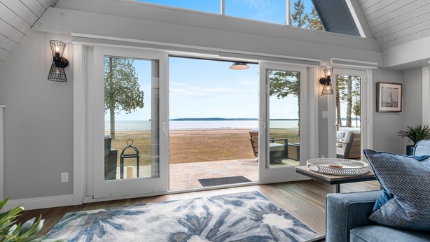 A modern interior with a large sliding glass door leading to a beach view, a cozy seating area with a patterned rug, and a dining table with a basket. - Moonlight Bay Retreat, Waterfront home near Cana Island. (Baileys Harbor)