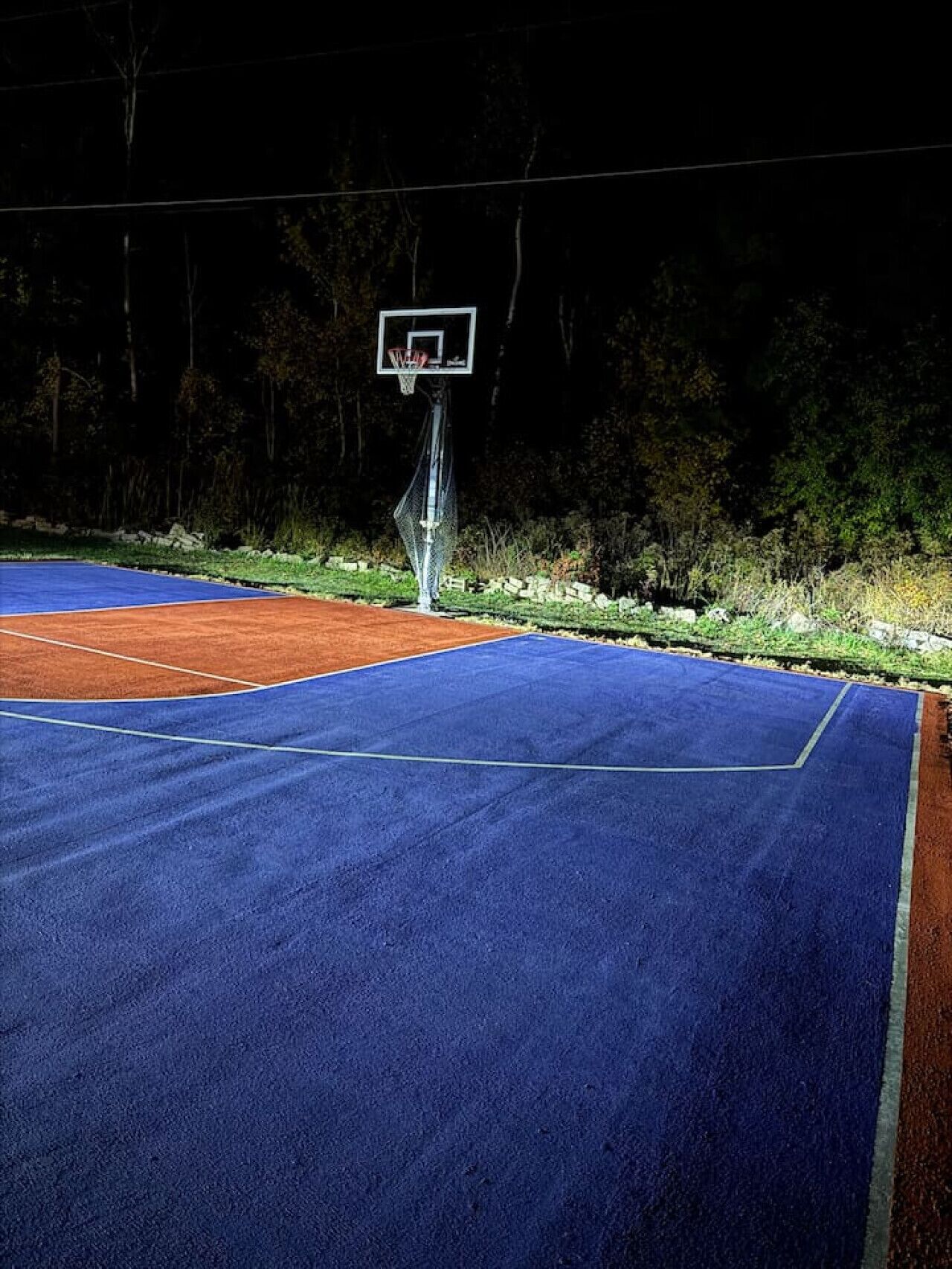 Un terrain de basket en plein air, éclairé la nuit, avec un panier et un panneau, entouré d’arbres et d’un mur de pierre.