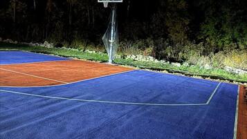 Un terrain de basket en plein air, éclairé la nuit, avec un panier et un panneau, entouré d’arbres et d’un mur de pierre.