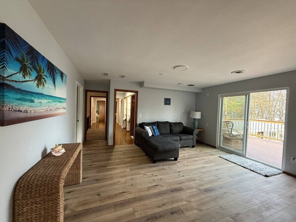 Un salón amplio con suelo de madera, un sofá gris oscuro, un banco y un cuadro de una playa.