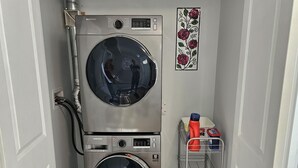 Un cuarto de lavandería con dos lavadoras de carga frontal apiladas, un estante metálico con productos de limpieza y un adorno floral en la pared.