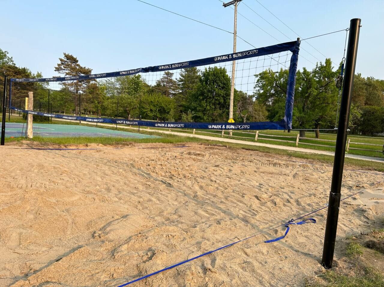 A volleyball net with "Park & Sun Sports" branding, set up on a sandy area with trees and a clear sky in the background.