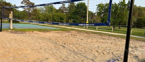 Ett volleybollnät med logotypen för "Park & Sun Sports", uppsatt på en sandig yta med träd och en klar himmel i bakgrunden.