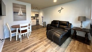 Un salon moderne avec un canapé en cuir noir, une table à manger avec des chaises, une cuisine en arrière-plan et un tableau accroché au mur.