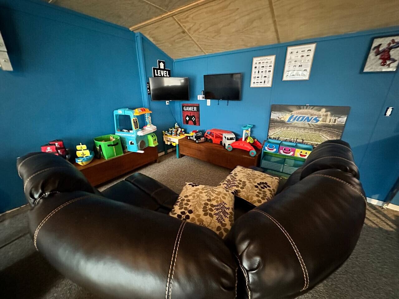 A room with a blue wall, a black leather couch, a gaming setup with toys and a TV, and a poster of 'Lions 1'.