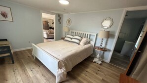 Une chambre à coucher avec un grand lit, un plancher en bois, une table de chevet avec une lampe et une porte donnant sur une autre pièce.