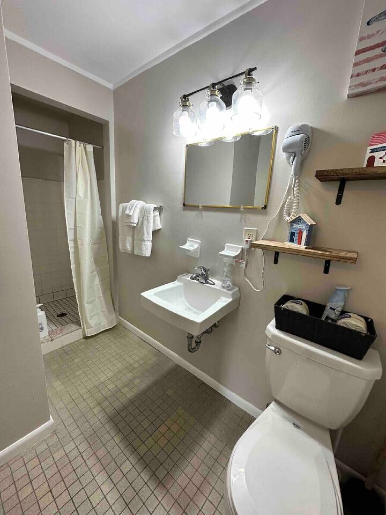 A bathroom with a sink, mirror, and toilet. There is a shower curtain, a hairdryer, and a shelf with toiletries.