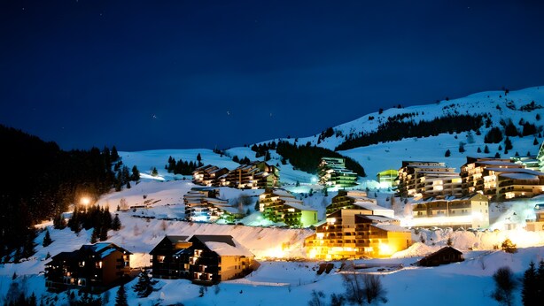 Un pueblo cubierto de nieve en la noche, con edificios iluminados y una pista de esquí al fondo.