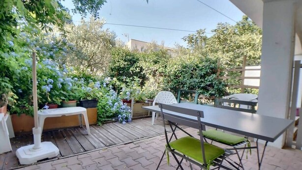 An outdoor patio area with a table and chairs, surrounded by potted plants and a wooden deck. - Appa L'olivier 18 rue Louis Pasteur (Saintes-Maries-de-la-Mer)