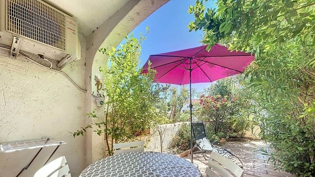 A round table with a patterned tablecloth, a pink umbrella, and white chairs in a garden setting. - Stud Résidence le trident 63 (Saintes-Maries-de-la-Mer)