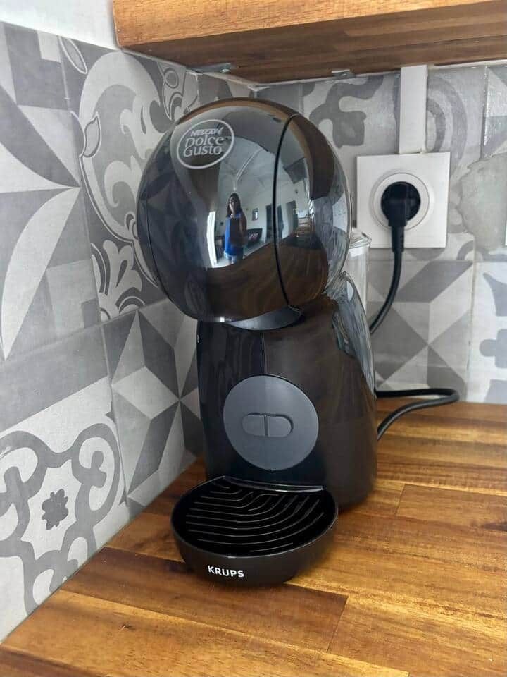 Une machine à café Krups posée sur un plan de travail en bois, avec un pare-éclaboussures à motifs.