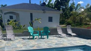 Une piscine rectangulaire à l’eau claire, entourée d’une terrasse carrelée. On aperçoit, au fond, des chaises longues et une petite table avec des chaises.