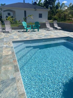 A rectangular swimming pool with clear water, surrounded by a tiled deck. There are lounge chairs and a small table with chairs in the background. - Gîte Mango-Passion Le Domaine Mannou (Sainte Rose)