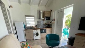 A modern living room with a beige sofa, colorful pillows, a teal armchair, and a small wooden coffee table. A kitchen with a microwave, oven, and washing machine is visible in the background. - Gîte Coco-orchidées Le Domaine Mannou (Sainte Rose)