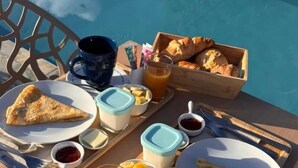 A breakfast tray with croissants, a slice of pizza, fruit, and beverages by a poolside table. - Gîte Coco-orchidées Le Domaine Mannou (Sainte Rose)