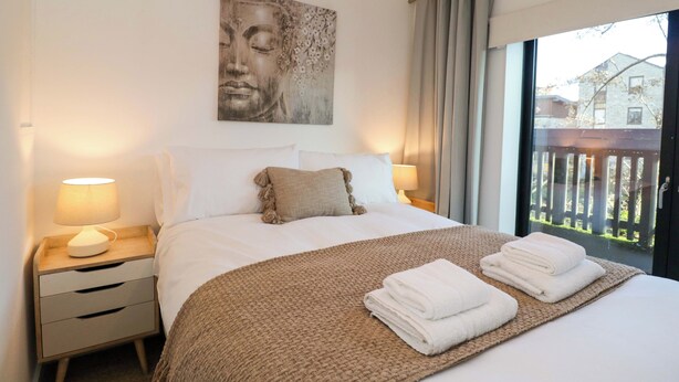 A neatly made bed with a brown blanket and two white pillows, a bedside lamp, and a painting on the wall. - 4 The Stoneyard (Bath)