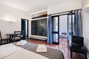 Habitación de hotel con dos camas, televisión, mesa de comedor, sillas, balcón con cortinas y un ventanal grande.