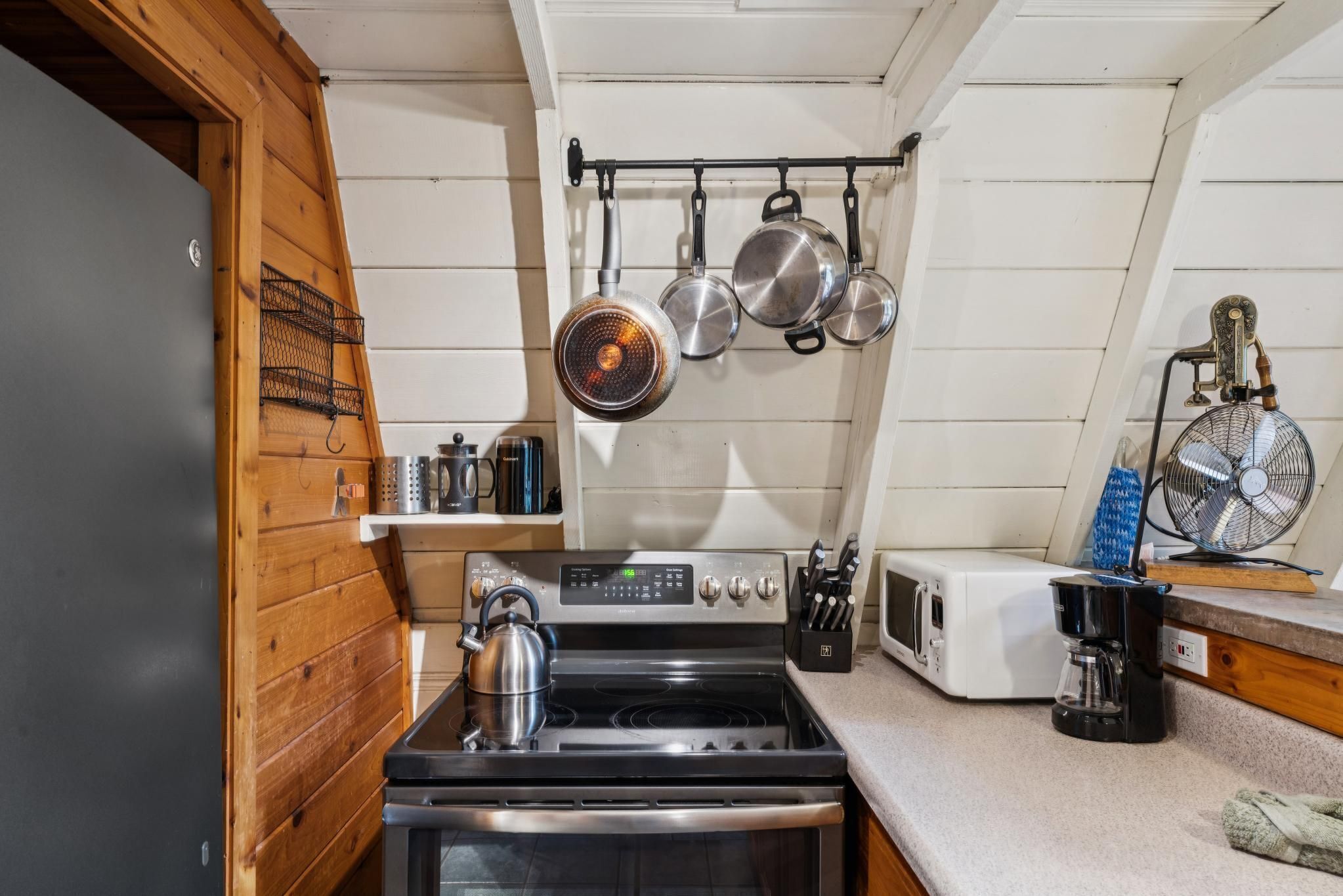 A kitchen with a stove, oven, hanging pots, a coffee maker, and a fan.