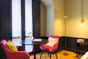 A room with two red chairs, a small round table with a laptop, a floor lamp, and a shelf with a bag.