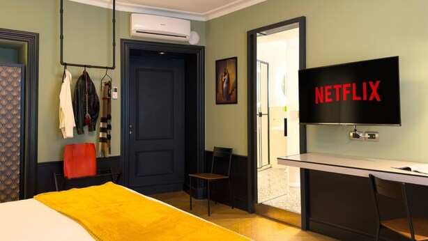 A hotel room with a bed, a desk with a TV displaying 'NETFLIX,' a coat rack, and an air conditioning unit. - GuestHost - In the heart of Brera, in a 20th-century Art Nouveau building on the 1st floor with elevator is the newly renovated double room with large bathroom, a cozy double bed, air conditioning, wifi, mini-bar, coffee maker, smart TV, bath linens, ill (Milan)