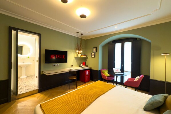 A hotel room with a bed, a desk with a laptop, a TV, a red trash bin, and a bathroom visible through an open door.