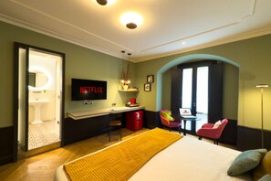 A hotel room with a bed, a desk with a laptop, a TV, a red trash bin, and a bathroom visible through an open door.