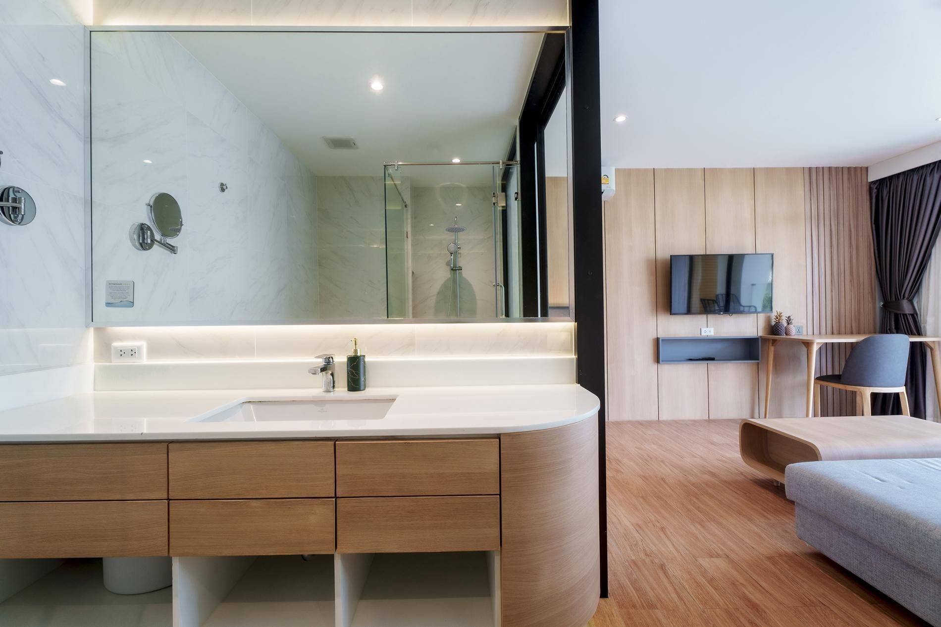 A modern bathroom with a large mirror, a sink, and a shower. There is a wooden vanity and a TV mounted on the wall.