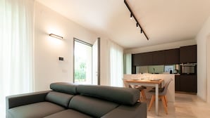 Ein modernes Wohnzimmer mit grauem Ecksofa, einem Essbereich mit Holztisch und -stühlen sowie einer Küche mit dunklen Schränken und einem eingebauten Backofen.
