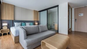 A modern hotel room with a grey sofa, a wooden coffee table, a bed with a headboard, and a glass door leading to a bathroom. - Wyndham Garden 1220 Modern Studio Next to Naitho (Sa Khu)