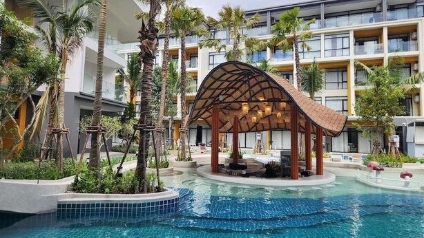 A resort pool area with a covered seating area, palm trees, and a modern building in the background. - Wyndham Garden 1220 Modern Studio Next to Naitho (Sa Khu)