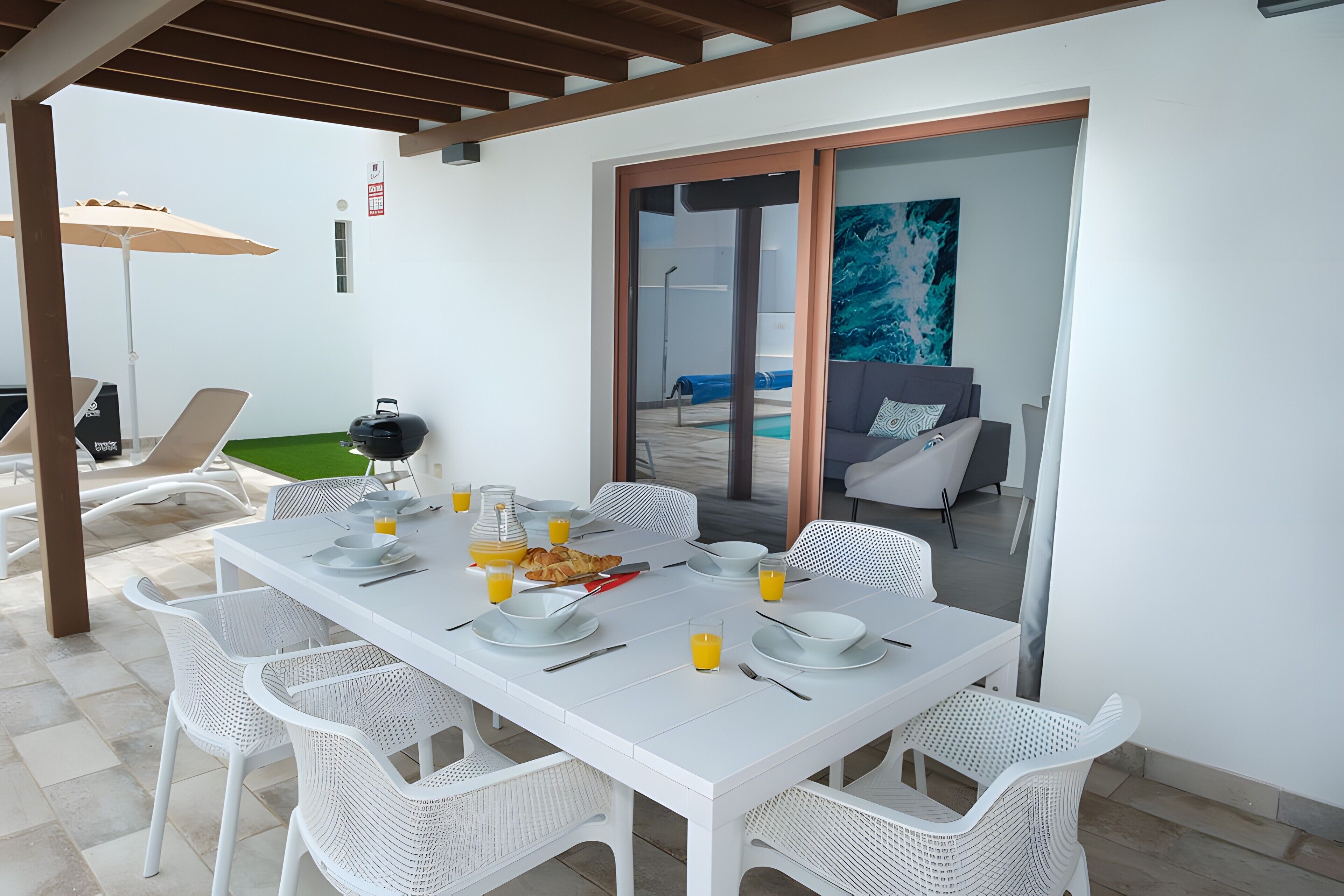 A dining area with a white table set for four, surrounded by white chairs, with a view of a pool and a sliding glass door.