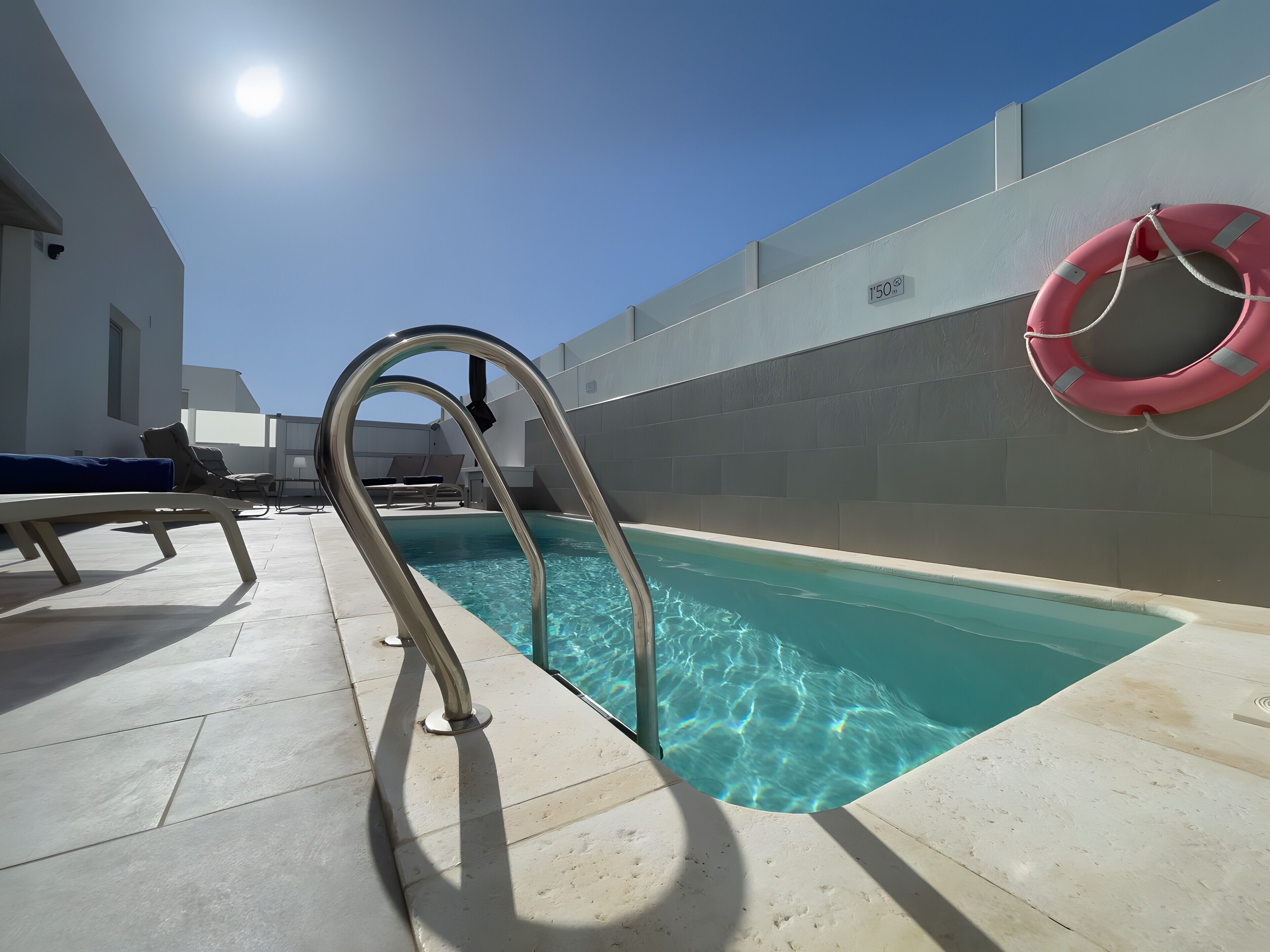 Una piscina con agua cristalina, una escalera metálica, sillas de descanso y un flotador en la pared.