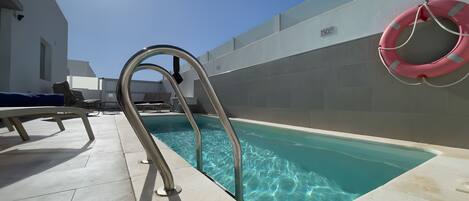 Una piscina con agua cristalina, una escalera metálica, sillas de descanso y un flotador en la pared.