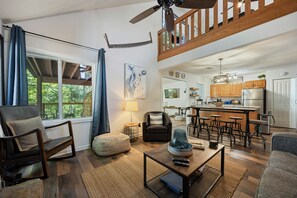 Un salón amplio con un gran ventanal, una zona de comedor con taburetes de bar, una cocina con armarios de madera y un ventilador de techo.