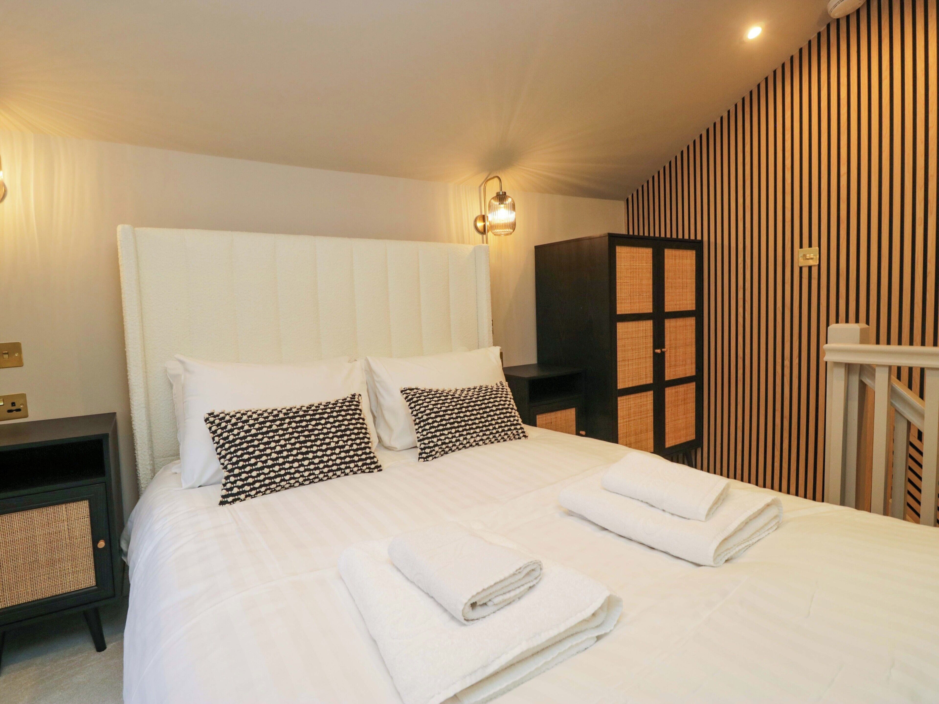 A neatly made bed with white linens and black and white patterned pillows, a bedside table with a lamp, and a television on a stand against a striped wall.