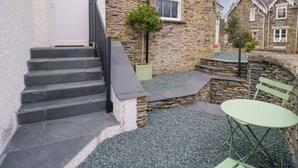 Un bâtiment en pierre avec une porte blanche, un petit escalier et une terrasse en gravier agrémentée d’une table et de chaises en métal vert.