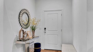 Un couloir avec une porte blanche, un miroir, une petite table ornée de quelques objets décoratifs et un banc.