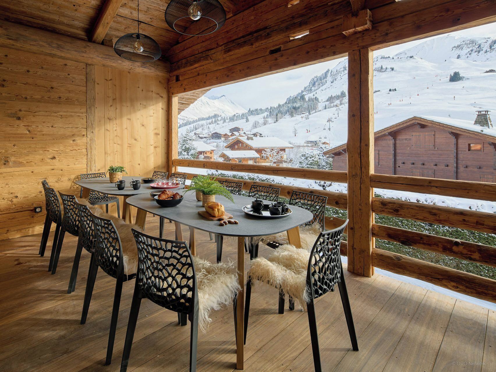 A wooden cabin with a dining area featuring a round table, chairs, and a view of a snowy mountain landscape.
