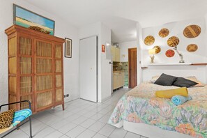 Ein Schlafzimmer mit einem Bett, einem Holzschrank, einem Sessel und einem durch eine offene Tür einsehbarer Küchenbereich.