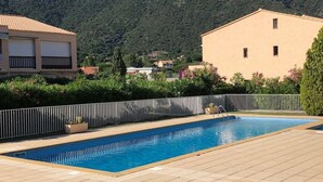 A swimming pool with a tiled deck, surrounded by a white fence, with buildings and a mountain in the background.