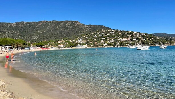 A beach with clear water, a mountain in the background, and boats.