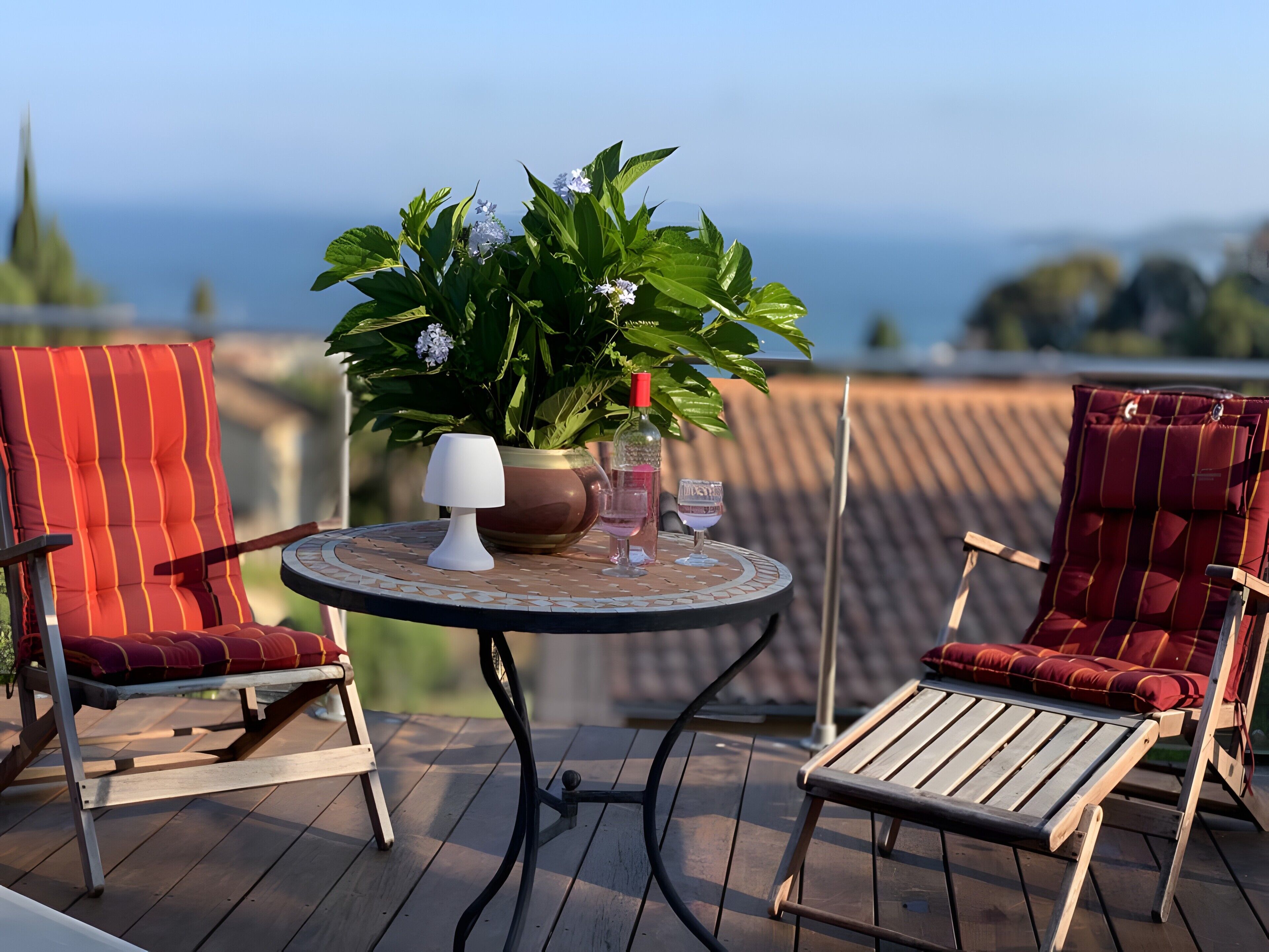A deck with two striped chairs, a table with a potted plant, a lamp, and two wine glasses.