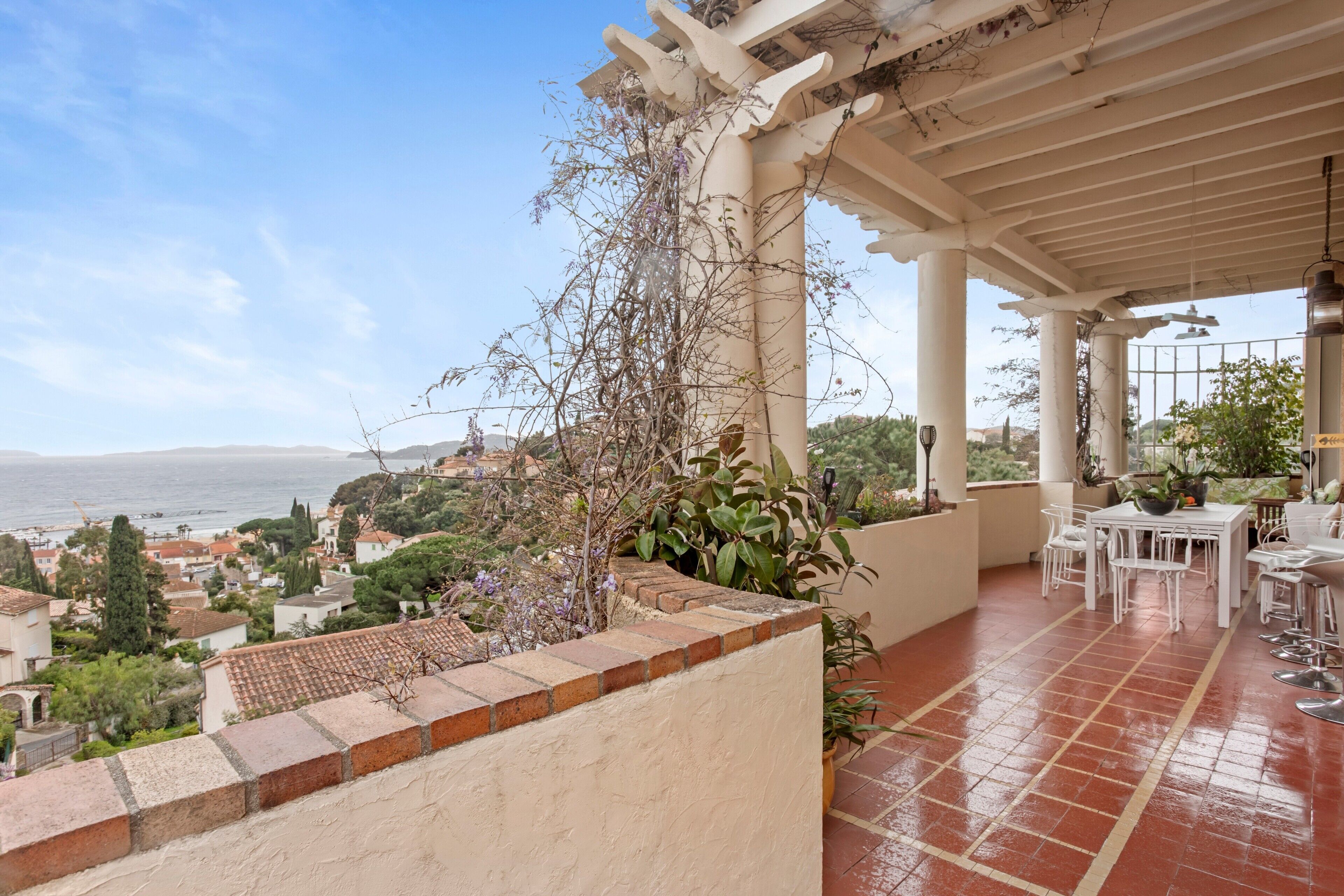 A covered outdoor area with a terrace, potted plants, and a view of the sea and buildings.
